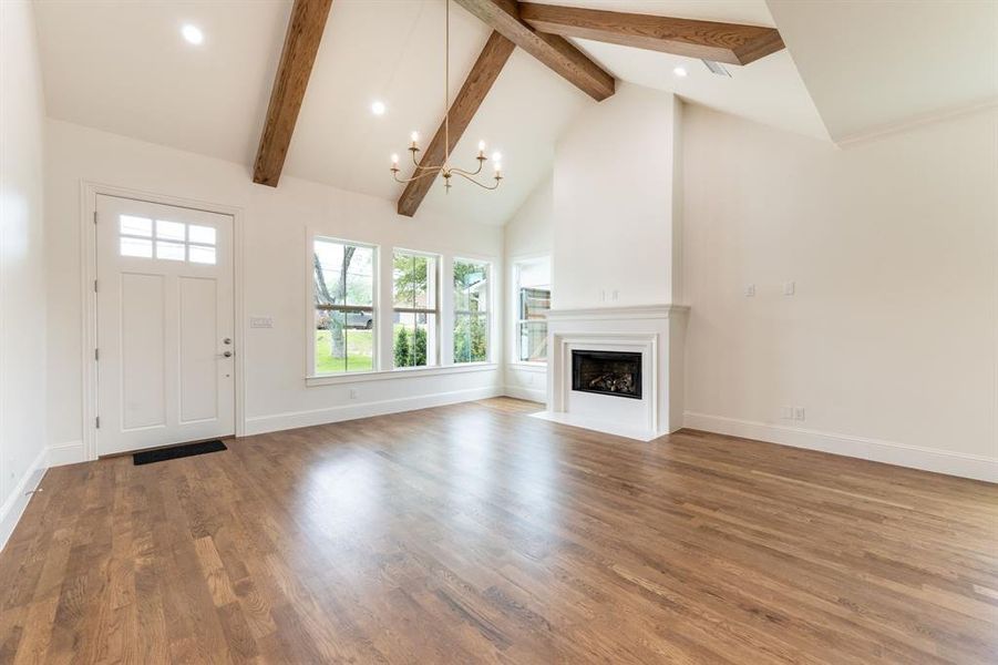 Vaulted ceiling with exposed wood beams, wood-finish flooring, white fireplace with mantle, three large windows, and a multi-pane entry door Vaulted ceiling with exposed wood beams, wood-finish flooring, white fireplace with mantle, three large windows, and a multi-pane entry door