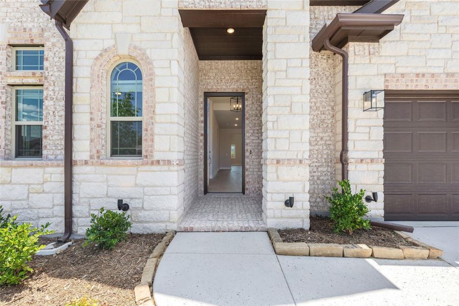 Home entryway with stone exterior, covered front door, brick walkway, and attached garage door visible to the right. Home entryway with stone exterior, covered front door, brick walkway, and attached garage door visible to the right.