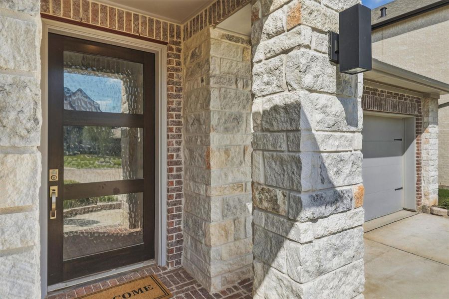 The inviting front entry showcases impressive stone and brick craftsmanship, sleek contemporary lighting, and an upgraded front door with decorative privacy glass and keyless entry. Clean architectural lines and quality finishes create a warm, welcoming arrival for family and guests alike.