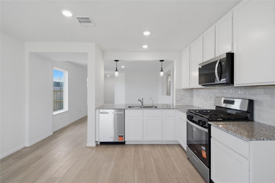 Kitchen boasts soaring ceilings, white stained Woodmont Dakota cabinets, Granite countertops in Iberian White, stainless steel Whirlpool appliances, a modern Daltile backsplash, recessed lighting, and a pantry—all overlooking the expansive family room.