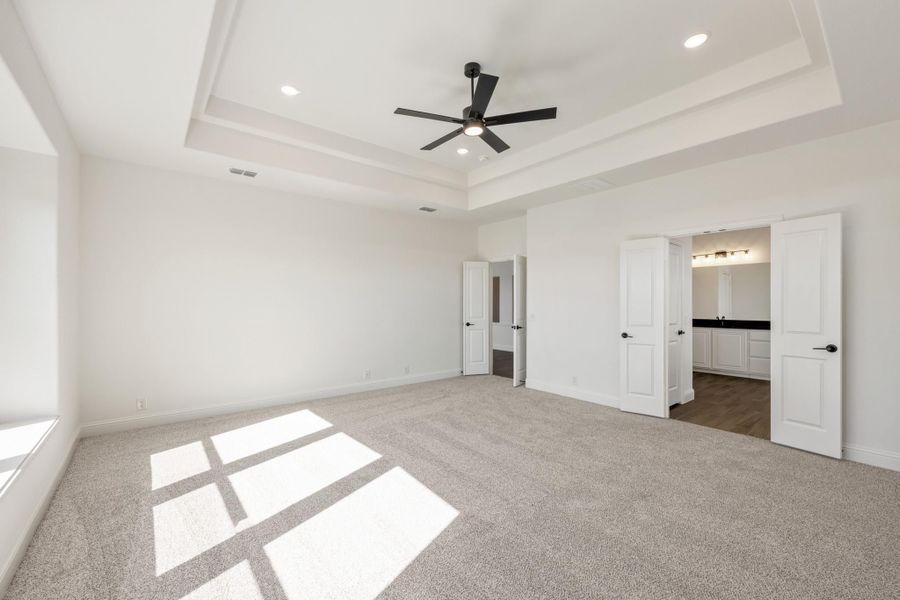 Spacious master bedroom with tray ceiling, ceiling fan, carpet flooring, and open ensuite bathroom doorway. Spacious master bedroom with tray ceiling, ceiling fan, carpet flooring, and open ensuite bathroom doorway.