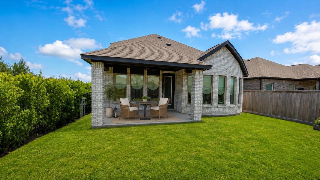 The backyard features an open green lawn bordered by a tall privacy hedge and a wooden fence. The back of the light brick home shows a covered concrete patio space.**This room has been virtually staged.