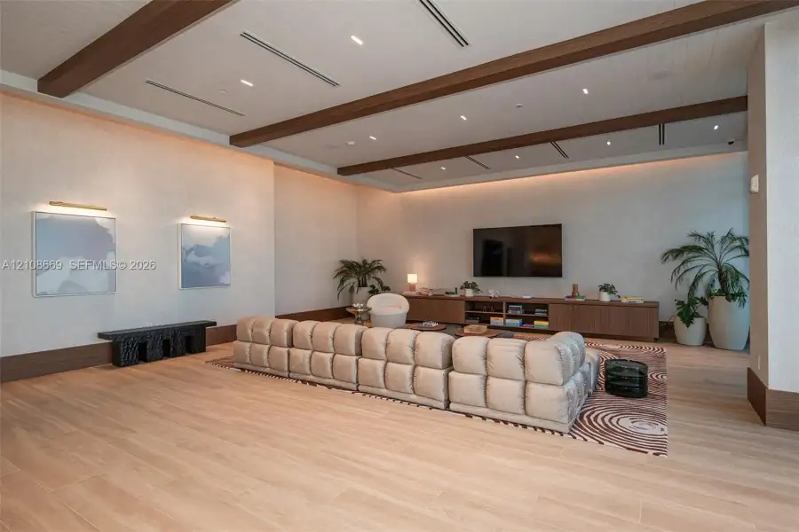 Large living area featuring wood-finish flooring, recessed ceiling lighting, decorative wood-beam ceiling accents, wall-integrated cove lighting, and custom built-in cabinetry