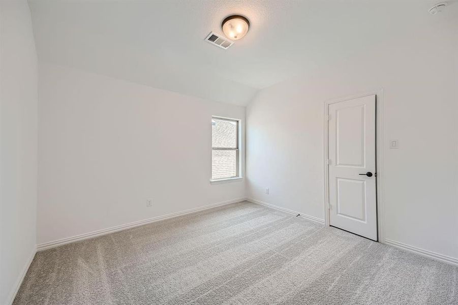 Spacious room featuring light gray patterned carpet, white walls, a single window with exterior brick detail, and a white paneled door with a matte black handle Spacious room featuring light gray patterned carpet, white walls, a single window with exterior brick detail, and a white paneled door with a matte black handle