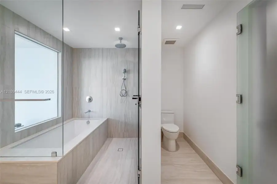 Bathroom featuring floor-to-ceiling stone-finish tile, glass-enclosed soaking tub with rain-style shower head, recessed lighting, and a private water closet