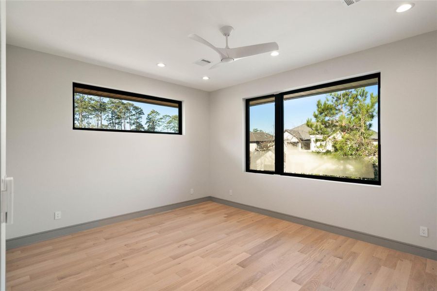 This bright, spacious room features large windows with scenic views, light wood flooring, recessed lighting, and a ceiling fan, offering a modern and inviting atmosphere.