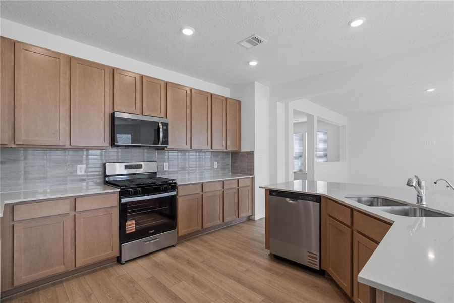 This spacious kitchen boasts soaring ceilings, Woodmont Dakota cabinets in a Barley stain, Silestone Linen Cream countertops, stainless steel Whirlpool appliances, a modern Daltile backsplash, recessed lighting, an extended breakfast bar countertop, and a pantry—all overlooking the expansive family room and breakfast nook.
