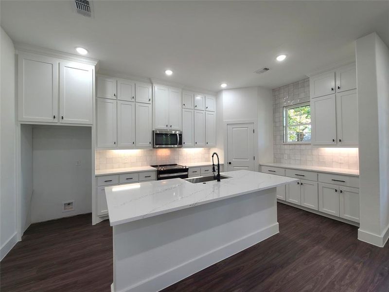 Kitchen featuring dark wood-type flooring, white cabinets, appliances with stainless steel finishes, and a kitchen island with sink