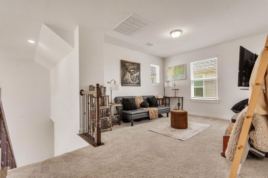 This cozy loft area features plush carpeting, ample natural light from two windows, a comfortable seating arrangement, and stylish decor. It’s perfect for relaxation or a small home office setup.