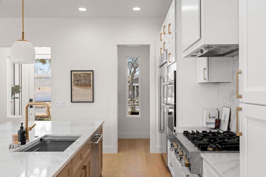 Dual tone kitchen with light stone counters, decorative light fixtures, light wood-style floors, stainless steel appliances, and ventilation hood