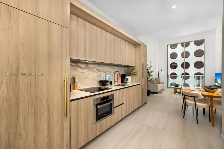 Contemporary kitchen featuring light wood-finish cabinetry, integrated appliances, a stone-pattern backsplash, solid surface countertops, and brushed gold-tone hardware