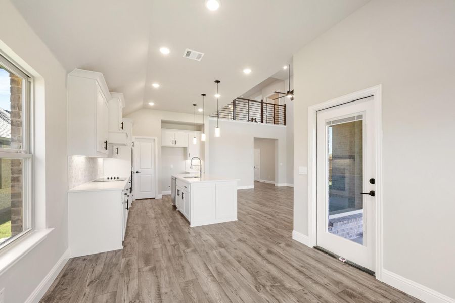 Open kitchen with white cabinets, center island, pendant lights, and wood floors under vaulted ceiling with upper loft railing. Open kitchen with white cabinets, center island, pendant lights, and wood floors under vaulted ceiling with upper loft railing.