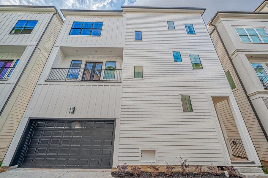 Modern three-story townhouse with a sleek facade, featuring a balcony and large windows. It includes an attached garage and a small landscaped area at the front.