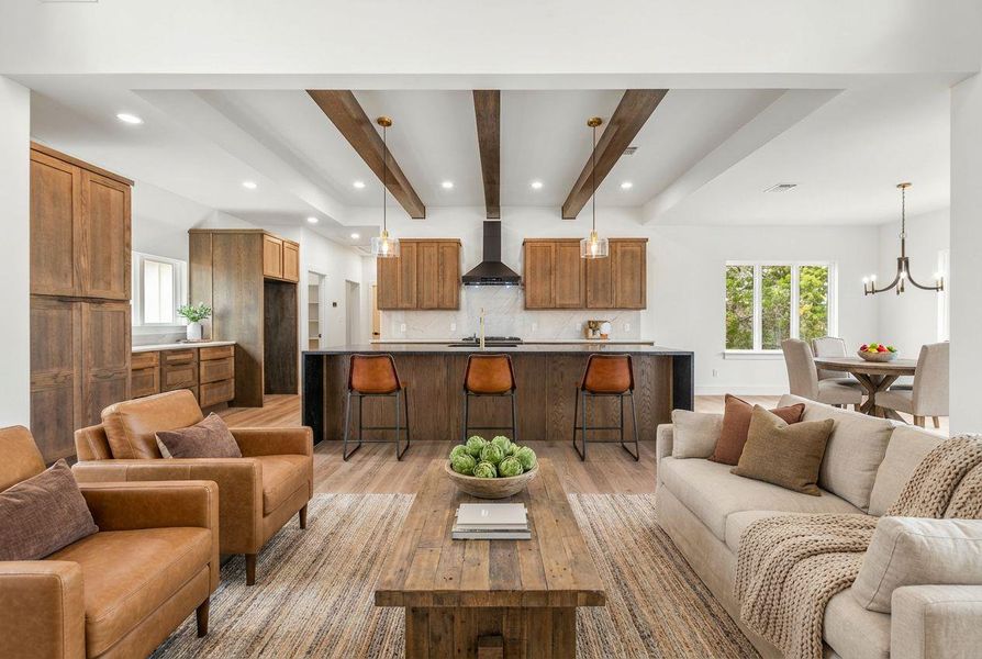 Striking white oak beams lead your gaze into the gourmet kitchen, featuring generous white oak cabinetry, all centered around a deluxe range hood and pendant lights illuminating the island.