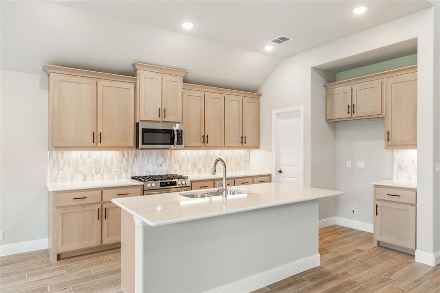 Kitchen with light wood cabinets, center island with sink, stainless steel appliances, and tile backsplash