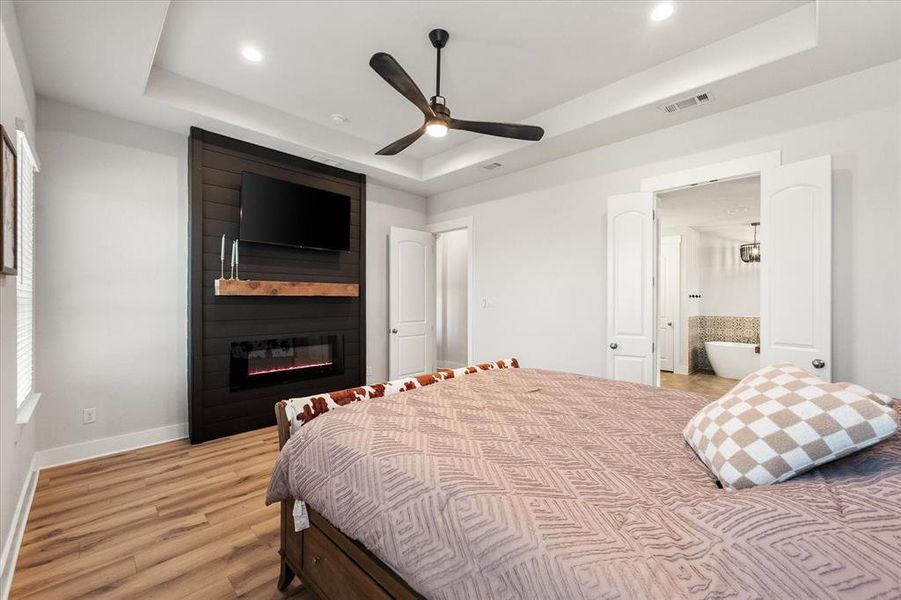 Bedroom featuring ensuite bathroom, a raised ceiling, light wood finished floors, a fireplace, and recessed lighting