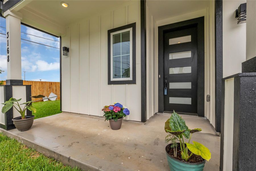 Step into modern elegance with this inviting front porch, featuring sleek black accents, vibrant potted plants, and a stylish glass-paneled door that welcomes you home.