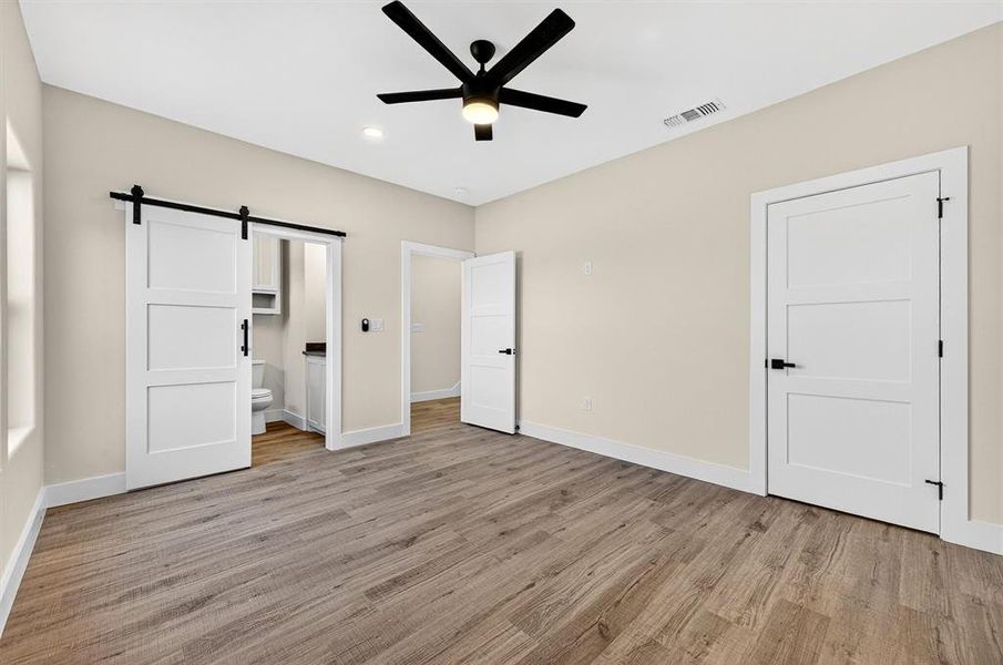 Unfurnished bedroom featuring a barn door, connected bathroom, light wood finished floors, and a ceiling fan