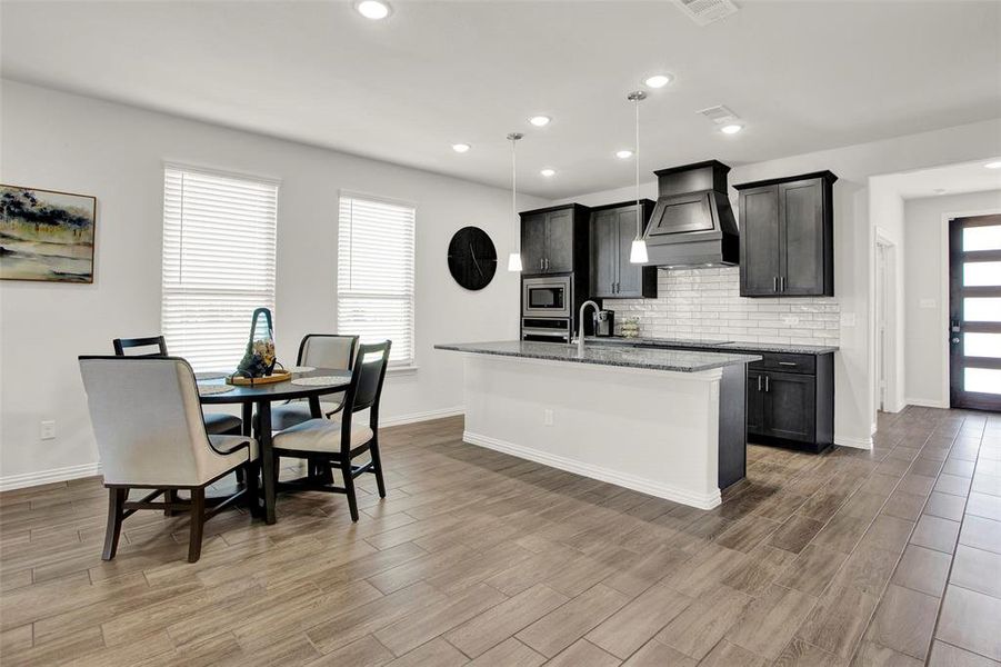 Kitchen with custom exhaust hood, hanging light fixtures, an island with sink, stainless steel microwave, and backsplash