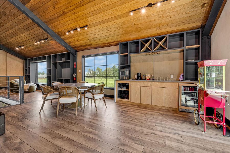 Wooden ceiling and floor, creating a warm atmosphere. It includes a stylish dining area with a round table and chairs, built-in shelves, and a counter with a wine rack. Large windows offer scenic views, enhancing the inviting space.