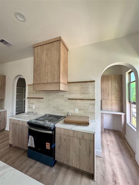 This modern kitchen features natural wood cabinetry and sleek countertops, with a new stove and stylish tile backsplash. It has an open layout with arched doorways leading to a bright, adjacent room.