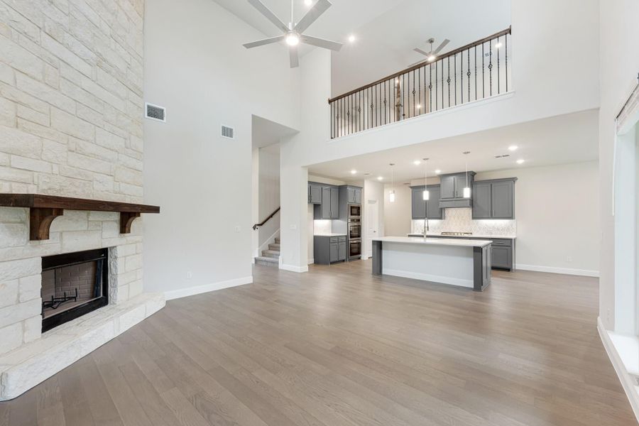 Open living area with stone fireplace, hardwood floors, and kitchen with gray cabinets and island visible in background Open living area with stone fireplace, hardwood floors, and kitchen with gray cabinets and island visible in background