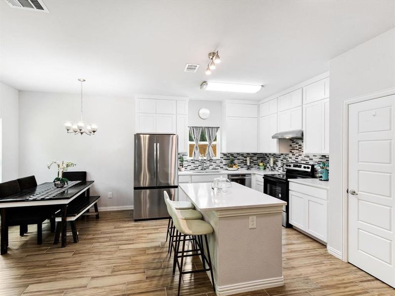 Modern kitchen featuring wood-finish flooring, white cabinetry, a large kitchen island with a light-toned countertop, a tile backsplash, and stainless steel appliances