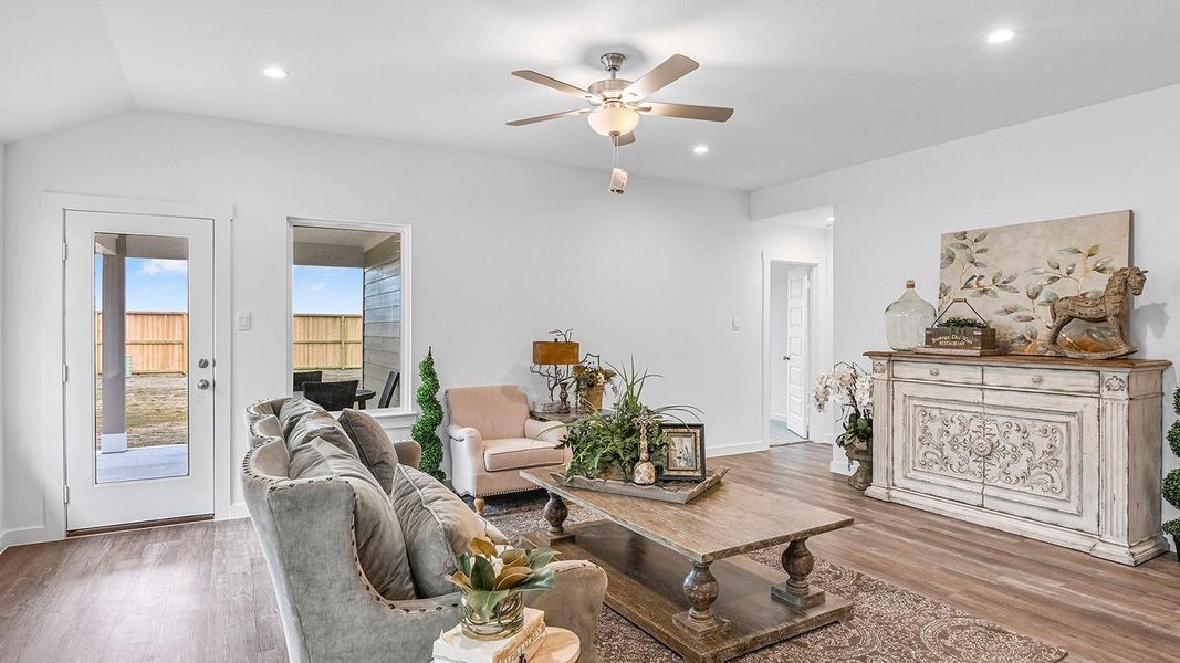 A cozy living room featuring large windows for natural lighting, wood flooring and a view to the kitchen and dining area.