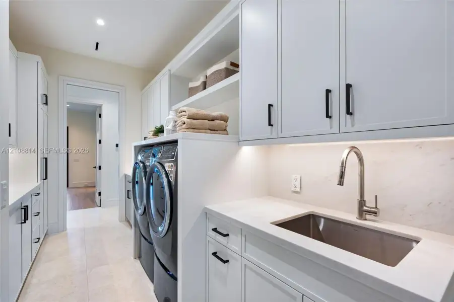 Laundry area featuring Shaker-style cabinetry, quartz countertops, a stainless steel undermount sink with a gooseneck faucet, light-toned stone-finish flooring, and recessed under-cabinet lighting Laundry area featuring Shaker-style cabinetry, quartz countertops, a stainless steel undermount sink with a gooseneck faucet, light-toned stone-finish flooring, and recessed under-cabinet lighting
