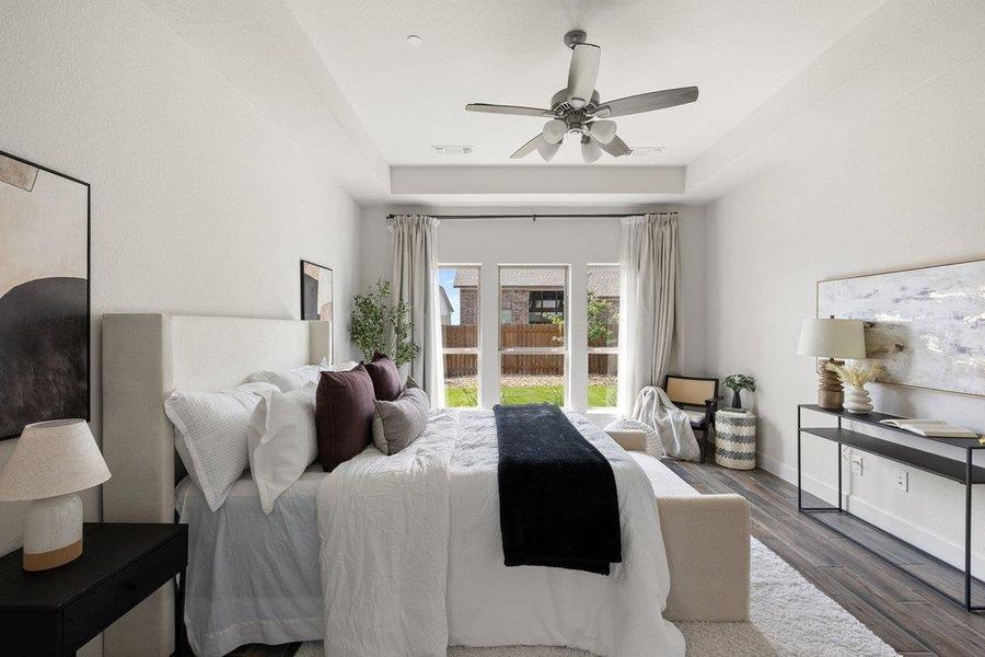 A tranquil primary bedroom with high ceilings, warm wood flooring, and a view of the green yard.
