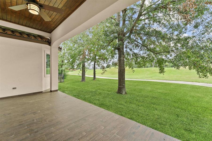 Spacious covered patio with a wood ceiling and ceiling fan, overlooking a lush, green lawn with mature trees and a scenic open space beyond.