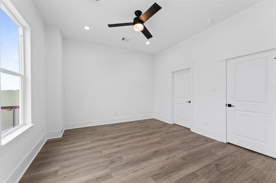 This photo shows a bright, modern room with white walls and wood-look flooring. It features a large window for natural light, a sleek ceiling fan, and two doors, likely leading to a closet and another room.