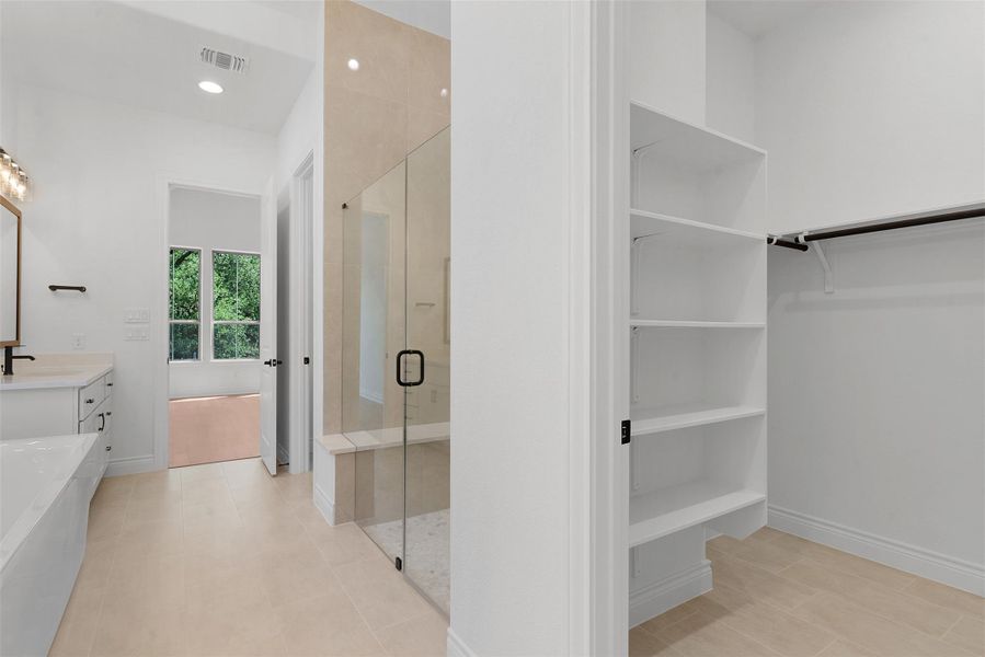 Spacious bathroom featuring light-colored tile flooring, a glass-enclosed shower with a bench, a white vanity with drawers, and a built-in closet with shelving and a hanging rod