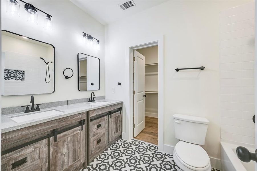 Bathroom featuring a dual vanity with light-toned countertops, wood-finish cabinetry with black hardware, and patterned floor tiling Bathroom featuring a dual vanity with light-toned countertops, wood-finish cabinetry with black hardware, and patterned floor tiling