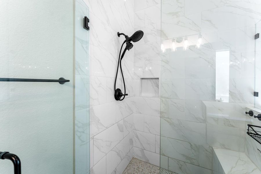 Shower enclosure featuring clear glass panels, matte black fixtures, white tile walls with grey veining, a built-in shower bench, and a recessed wall niche
