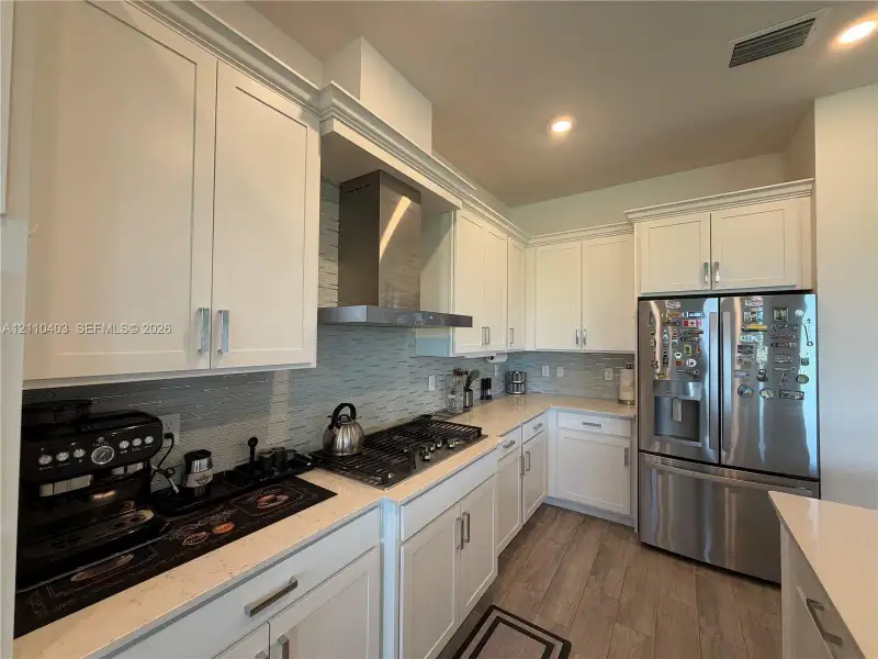 Kitchen featuring white shaker-style cabinetry, stainless steel appliances, a gas cooktop with a professional-style range hood, light-toned stone-finish countertops, and wood-finish flooring