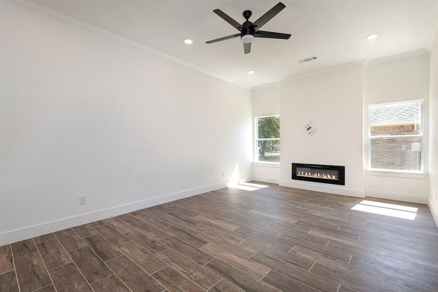 Unfurnished living room featuring a glass covered fireplace, ornamental molding, a ceiling fan, visible vents, and dark wood finished floors
