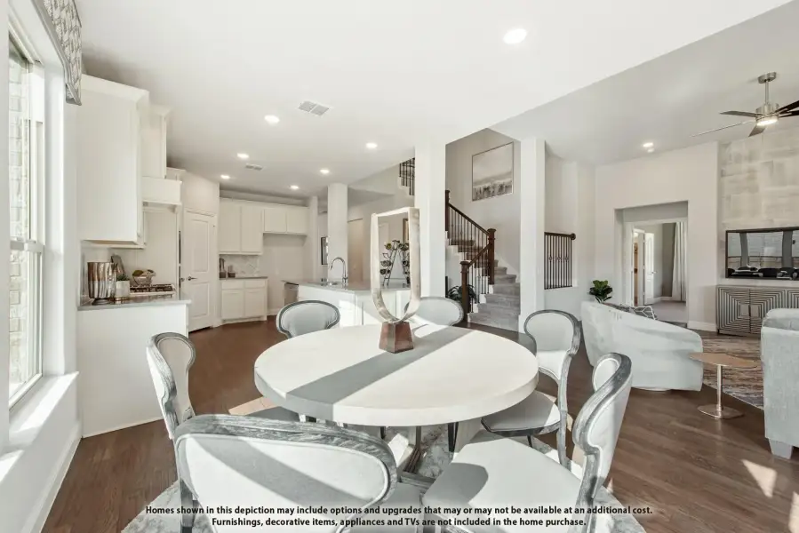 Open-concept dining area with round white table, hardwood floors, and kitchen visible in background near staircase.