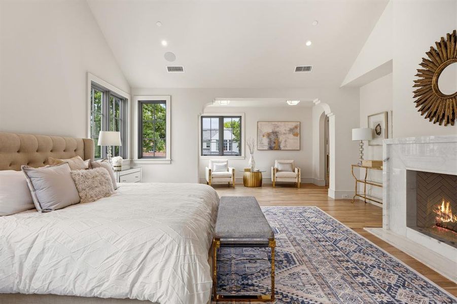 Spacious bedroom featuring vaulted ceilings, light wood-finish flooring, a white marble fireplace with a herringbone pattern firebox, and multiple windows with dark frames Spacious bedroom featuring vaulted ceilings, light wood-finish flooring, a white marble fireplace with a herringbone pattern firebox, and multiple windows with dark frames