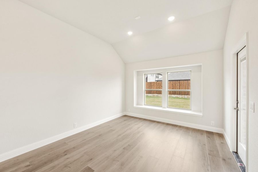Empty room with vaulted ceiling, light hardwood floors, and a large window overlooking a backyard fence.