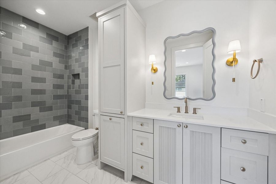 Chic secondary bath pairs a crisp white vanity with fluted cabinet detailing and warm brass fixtures for a refined finish. Glossy gray tile surround adds depth to the tub and shower, creating a polished yet playful design moment.