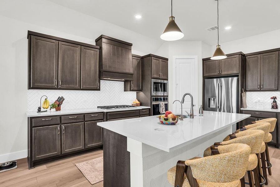 Kitchen featuring a substantial island with breakfast bar, undermount sink with gooseneck brushed nickel faucet, wood-finish flooring, dark wood cabinetry, a white herringbone tile backsplash, walk-in pantry and stainless steel appliances including a propane gas cooktop, electric built-in oven and microwave open to living and breakfast rooms