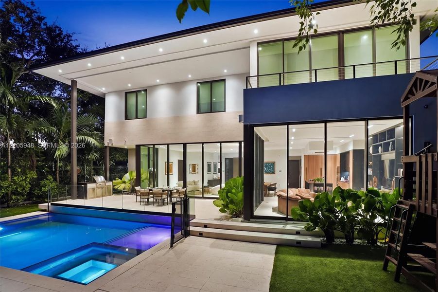Contemporary two-story residence featuring expansive floor-to-ceiling glass sliding doors, a built-in outdoor kitchen, a private swimming pool with integrated spa, and a second-level balcony with metal railing