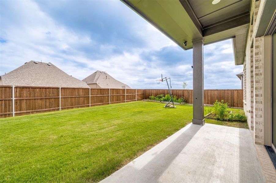 Expansive backyard featuring a concrete patio, a covered porch with recessed lighting, a lush green lawn, a wood privacy fence, and planted garden beds