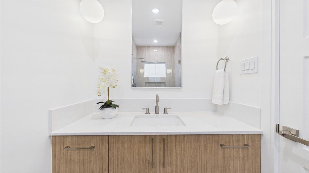 This bathroom features a modern vanity with a sleek white countertop and integrated sink. It has contemporary fixtures, a large mirror, and soft lighting. The wooden cabinetry adds warmth, while a towel ring and decorative plant enhance the space.