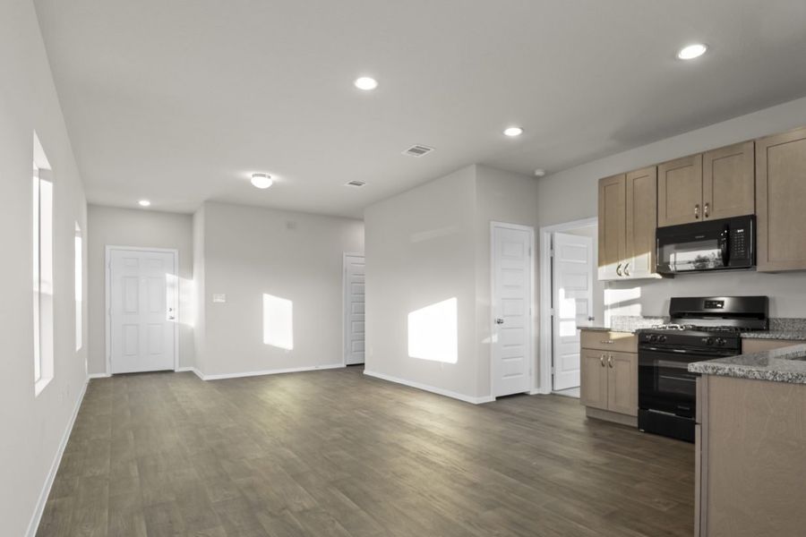 Photo of home living areas looking from the kitchen to the front door. White walls, wood-look floors, and light brown kitchen cabinets. Two windows on the left and kitchen and doors on the right.