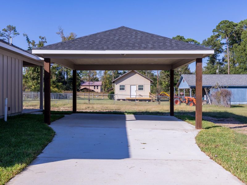 This photo shows a covered carport with a solid roof and wooden posts, set on a concrete driveway. The area is surrounded by grassy lawns and neighboring structures, offering a spacious and practical outdoor feature.