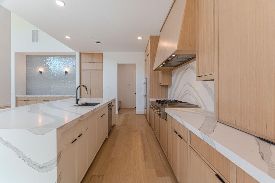 This kitchen features a modern design with light wood cabinetry and sleek white countertops. It includes a large island with a sink and ample workspace. The room is well-lit with recessed lighting, and the backsplash adds a touch of elegance. Perfect for a contemporary home.