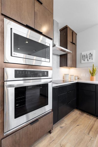 Kitchen featuring appliances with stainless steel finishes, wall chimney range hood, modern cabinets, light wood-style floors, and tasteful backsplash