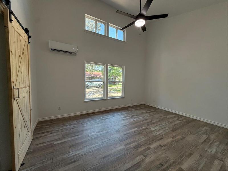 Unfurnished bedroom with a barn door, a towering ceiling, dark wood-style floors, ceiling fan, and a wall unit AC